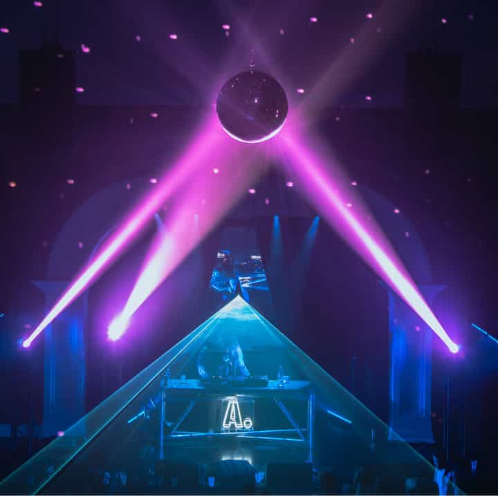Inside of a club with a disco ball above a crowd, a DJ on stage, and strobe lights illuminating the room.