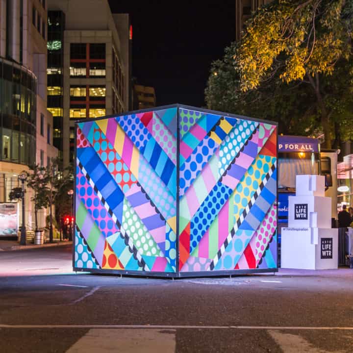 A square ratio shot of a large illuminated cube in the street decorated with art at night.
