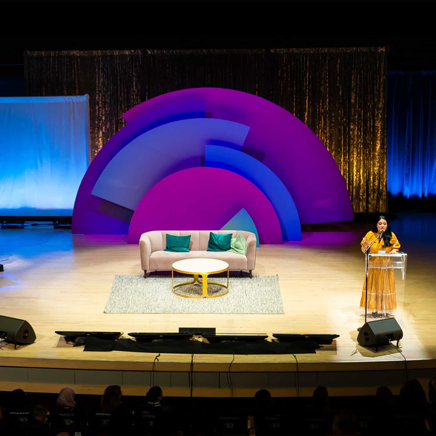 A high view of the Move the Dial stage with a woman in a yellow dress talking at a podium to the right.