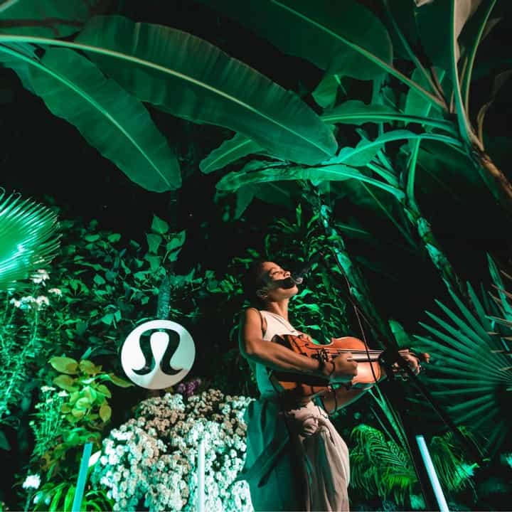 A low view of a woman playing violin in front of a lush plant background.